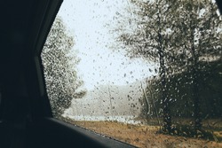 Rain drops through a car window in the middle of the dark dense forest, rainy and cloudy day in the mountains foggy background including a lake and tall trees, nostalgic mood