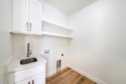 Empty white laundry room with top storage, laundry connections and dryer vent on the wall. There is a single vanity deep stainless sink beside laundry water outlet on the wall with electrical plug.