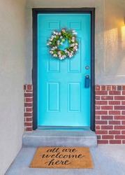 Vertical House exterior with red bricks and cyan front door with lockbox and wreath. Entrance of a house with statement doormat and open wall lamp lighting.