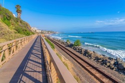 View of the train tracks and ocean from the bridge at San Clemente, California. There is a view of a mountain slope with wild shrubs and buildings near the pier against the blue sky at the back.