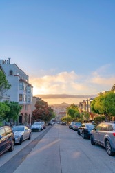 Residential area with roadside parking space and a view of sunset sky in San Francisco, CA. There are parked vehicles on both sides of the straight road with trees near the houses on the side.