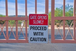 Gate opens slowly Proceed with caution signage on a metal gate at Tucson, Arizona. Close-up of a signage posted on the iron gate against the view of a concrete road and plants at the back.