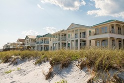 Destin, Florida- White sand dunes with grasses at the front of three-storey houses at the beach. Beach houses facade with balconies against the sky.