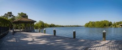 Wooden gazebo on a wood deck with piles at Milton, Florida. There is a gazebo on the left against the views of a waterfront and trees on the land area against the blue sky in panorama.