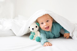 Little cute baby boy, child in knitted sweater, holding knitted toy, smiling happily at camera in white sunny, bright bedroom