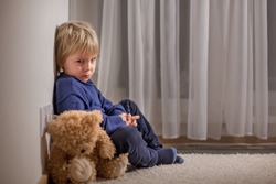 Angry little toddler child, blond boy, sitting in corner with teddy bear, punished for mischief
