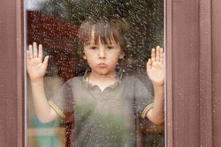 Little boy behind the window in the rain, looking sad