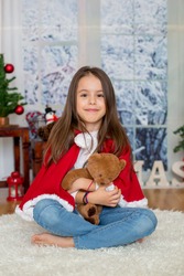 Happy child with toy sitting in front of  Christmas tree. Kid dressed in Santa Claus dress. Girl having fun at home
