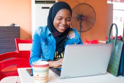 young nigerian muslim woman sitting alone in a cafe smiling while using her laptop