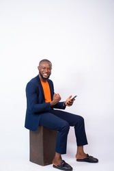 handsome young black man wearing a suit using his phone feeling excited and happy, sitting against a white plain background