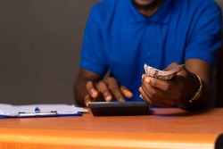 young african man sitting at a desk holding some money and doing some calculations
