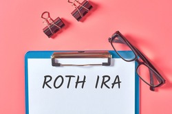 Clipboard with words roth ira near stationery items on pink desk. Business concept