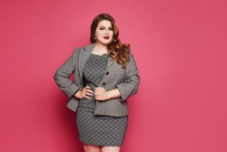 Charming plus size businesswoman wearing a grey jacket and dress looking at the camera and standing at pink background, isolated. XXL model in official outfit posing over pink