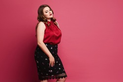 Young plus-size model girl in red satin blouse and skirt posing over pink background. Pensive chubby woman in modish outfit over pink background