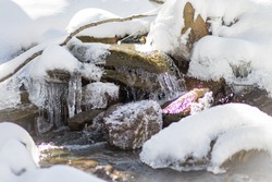 An icy and snowy stream. The snow and ice are melting in the springtime in Vermont. The water has formed icicles off the green and yellow rocks. The water is flowing slowly along the snow bank.