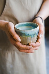 person in the apron holding handmade clay and turquoise cup of tea
