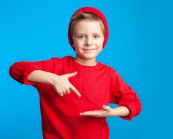 A beautiful 6-year-old boy in red on a blue background points his finger at the palm of his hand. Happy child. Portrait of a cute guy in bright fashionable clothes.