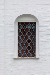 A narrow vertical window with bars in the white brick wall of the fortress. Vertical.