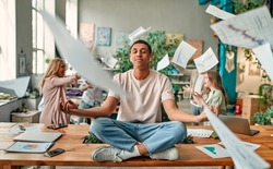 Keep calm and no stress! Young African-American man is sitting in lotus position while his colleagues are arguing nearby. Multiracial people working together in modern office.