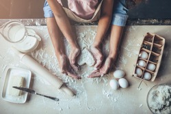 Cropped image of attractive young woman and her little cute daughter are cooking on kitchen. Having fun together while making cakes and cookies. Top view of mom with daughter making dough heart.