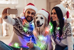 Merry Christmas and Happy New Year! Happy couple with dog labrador retriever waiting for the New Year in Santa Claus hats while sitting near beautiful Christmas tree at home. Smiling and making selfie