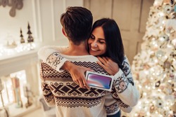 Merry Christmas and Happy New Year! Beautiful couple is waiting for the New Year together while hugging with gift in hand near beautiful Christmas tree at home. Modern smart phone.