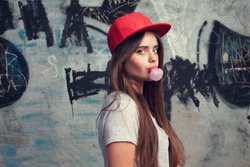 trendy beautiful long haired young model posing on graffiti background. Blow bubblegum. red cap. grey t-shirt.