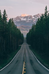 An open road leads to the Grand Teton's mountain range, rising in the distance beyond a thick pine forest. The last rays of sunlight shine on the mountain. Photo shot vertically to include more road. 