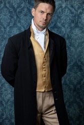 A handsome Regency gentleman wearing a gold waistcoat and black jacket and standing in a room with blue wallpaper and a wooden floor