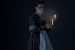A working class Victorian woman wearing a dark green checked bodice and skirt and holding a candle in a darkened room