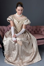 A Victorian woman wearing a gold ballgown and sitting on a pink, antique sofa against a studio backdrop