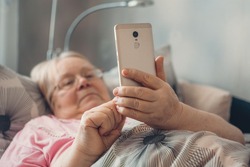 Social isolation, loneliness in older people, health risks. Age-group risk for coronavirus. Lonely elderly woman with smartphone lies in bed at home.