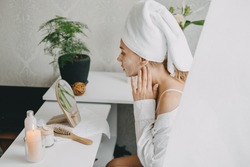 Young woman in white towel chilling in bedroom and making clay facial mask near mirror. Girl doing beauty treatment and relaxing at home. Morning skin care beauty routine, self care.