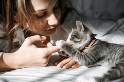 Candid portrait of Young woman is resting with kitten on the bed at home one sunny day. Girl play with outbred kitten.
