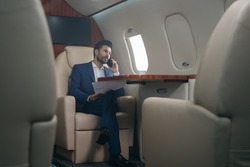 Handsome pensive middle eastern businessman talking on mobile phone, holding financial report working with documents sitting in plane. Confident entrepreneur flying private jet, successful business