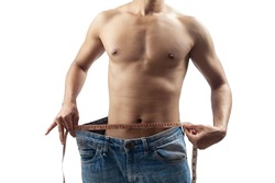 A slim young guy wearing a big jeans and measuring his waist by a tape measure over grey background.