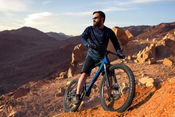 Cyclist in shorts and jersey on a modern carbon hardtail bike with an air suspension fork rides off-road on the orange-red hills at sunset evening in summer	