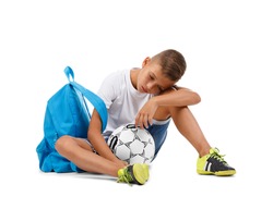 A cute boy with a bright satchel and a soccer ball isolated on a white background. A sitting child. Tired kid. Copy space.