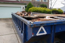 Long blue dumpster full of wood and other debris in the driveway in front of a house in the suburbs