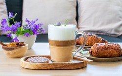 Beautiful cup of coffee latte macchiato on wooden table with sweet pasrty, croissant, cofee bread and purple flowers on wooden table in the living room, pastel tone 