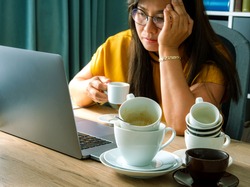 A piles of used coffee cups on desk front of an overtired Asian woman holding a cup of coffee sitting working frustrated exhausted looking at laptop screen. Caffeine addicted bad lifestyle concept. 