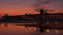 Pier silhouette Oceanside California USA. Pacific ocean tide tropical beach. Summertime gloaming atmosphere. Purple aesthetic gradient, calm twilight sky, pink violet dusk. Lights reflection in water.