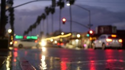Cars lights reflection on road in rainy weather. Rain drops on wet asphalt of city street in USA, water raindrops falling on sidewalk. Palm trees and rainfall, twilight dusk. Ocean Beach, California.