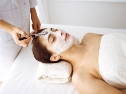Beautiful brunette woman enjoying applying cosmetic mask with closed eyes. Relaxing treatment in medicine and spa center concepts