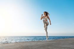 Young motivated happy sporty female runner fitness instructor, jumping, excercise near sea enjoying fresh morning air training, smiling facing sunlight, workout wearing activewear, running in quay