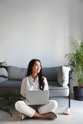 Vertical image of ambitious freelance girl sitting at home with laptop and looking outside window, smiling. Woman working on computer in living room.