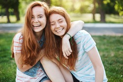 Wanna hug her forever. Portrait of happy and carefree two twin sisters with natural red hair and freckles, laughing out loud and cuddling, fooling around while resting in park on fresh green grass.
