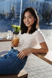 Vertical portrait of happy modern girl sitting in cafe near window and lean on table, drinking ice latte on hot summer day, smiling at camera.