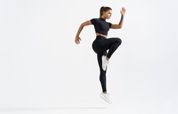 Dynamic movement of young and healthy sports woman doing jump exercising in sportswear. Fitness female in sport costume raising leg, running on white background.