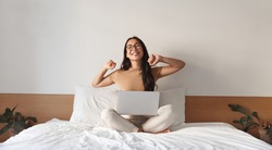 Image of satisfied young woman stretching arms, finish working on laptop. Asian girl sitting with computer in bed, wearing glasses, studying at home, e-learning and remote freelance job concept
